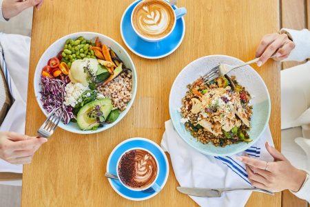 two people sitting at a wooden table eating bowls of food and drinking lattes out of blue ceramic mugs