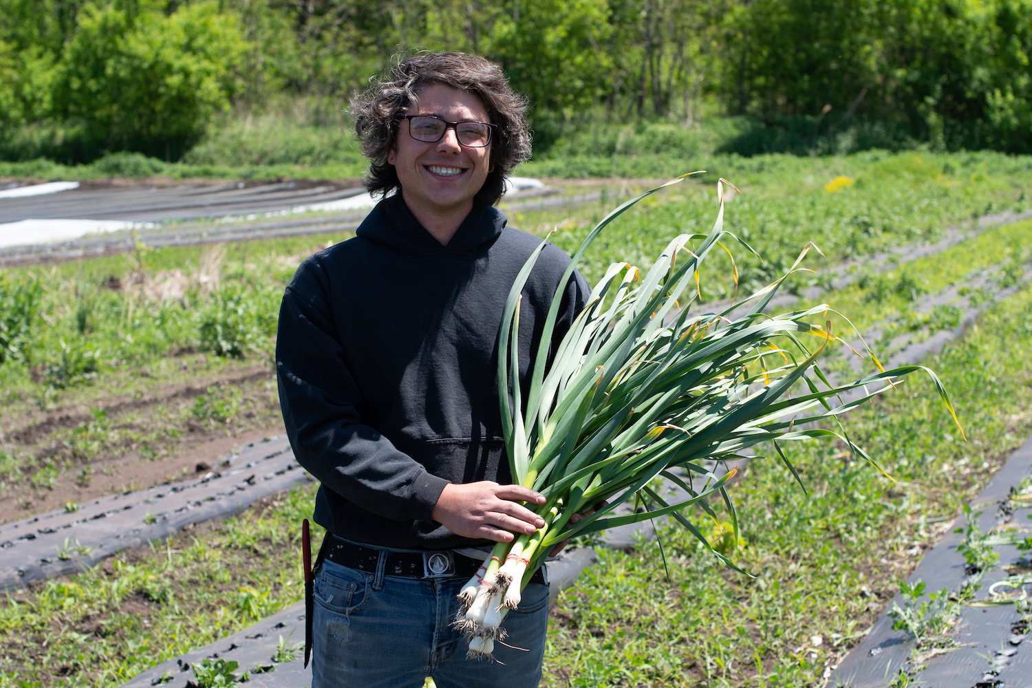 a man with curly hair and glasses standing in a garden holding green garlic