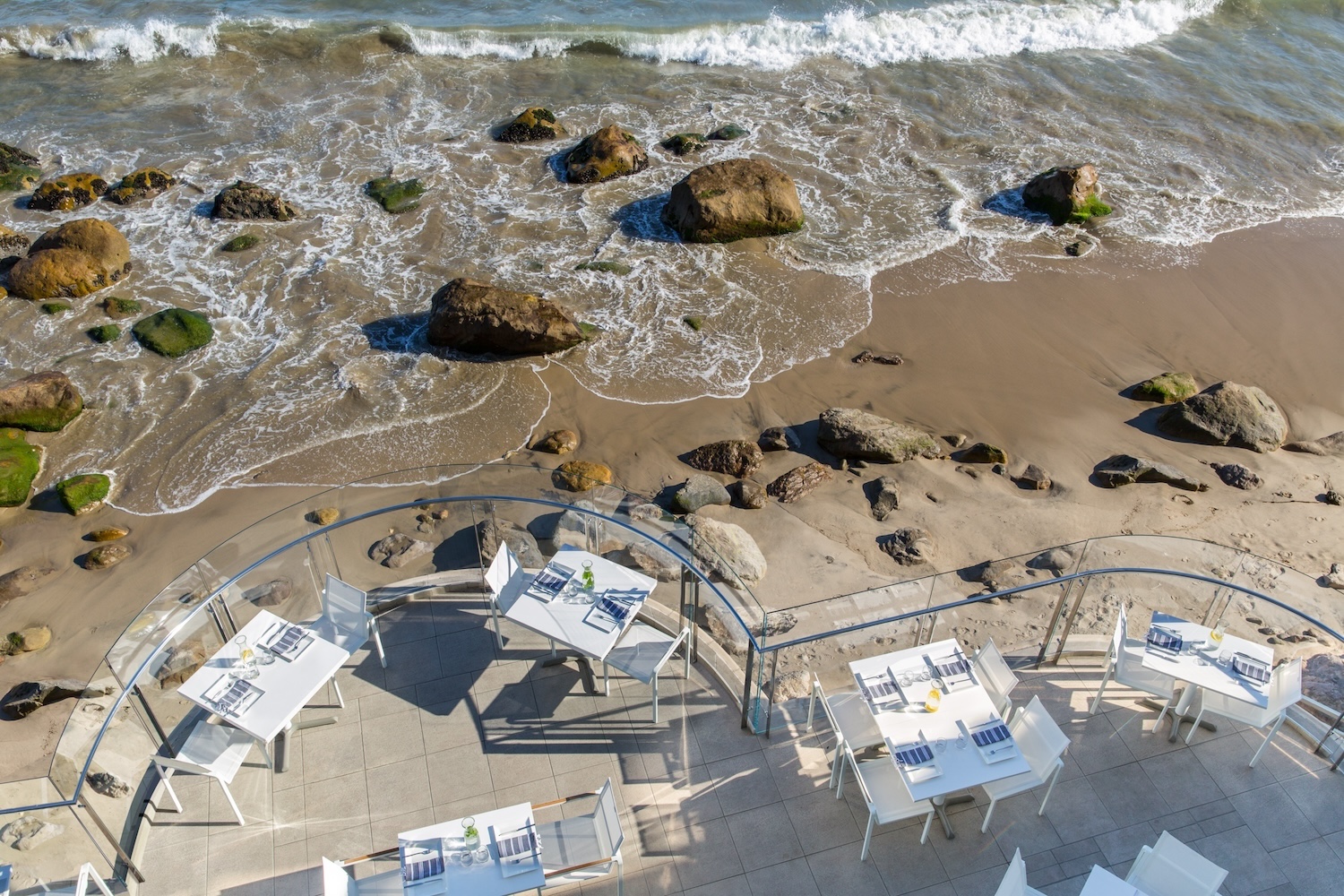 rocks, ocean shore, sand, terrace with white tables and chairs