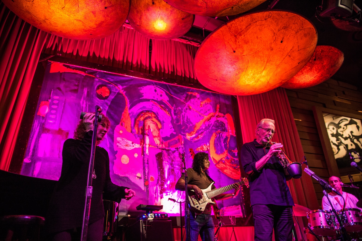 man playing trumpet and others playing instruments on stage, pink and red lights