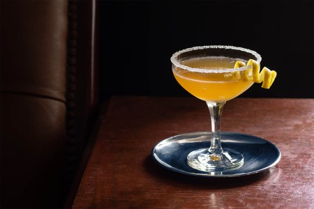 A chilled Sidecar cocktail in a coupe glass with a sugared rim on a dark wood table next to a leather seat. The Sidecar is gaining in popularity in top bars.