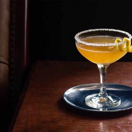 A chilled Sidecar cocktail in a coupe glass with a sugared rim on a dark wood table next to a leather seat. The Sidecar is gaining in popularity in top bars.