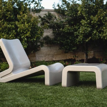 A grey lounge chair and ottoman sitting on grass in a backyard with a wall behind them