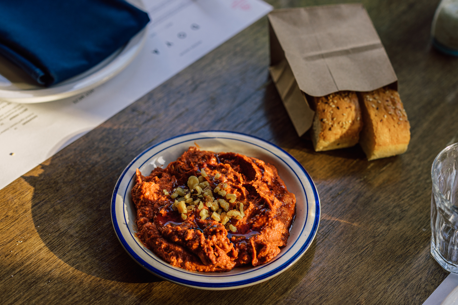 a dish of muhamarra on a wooden table next to a bag of bread