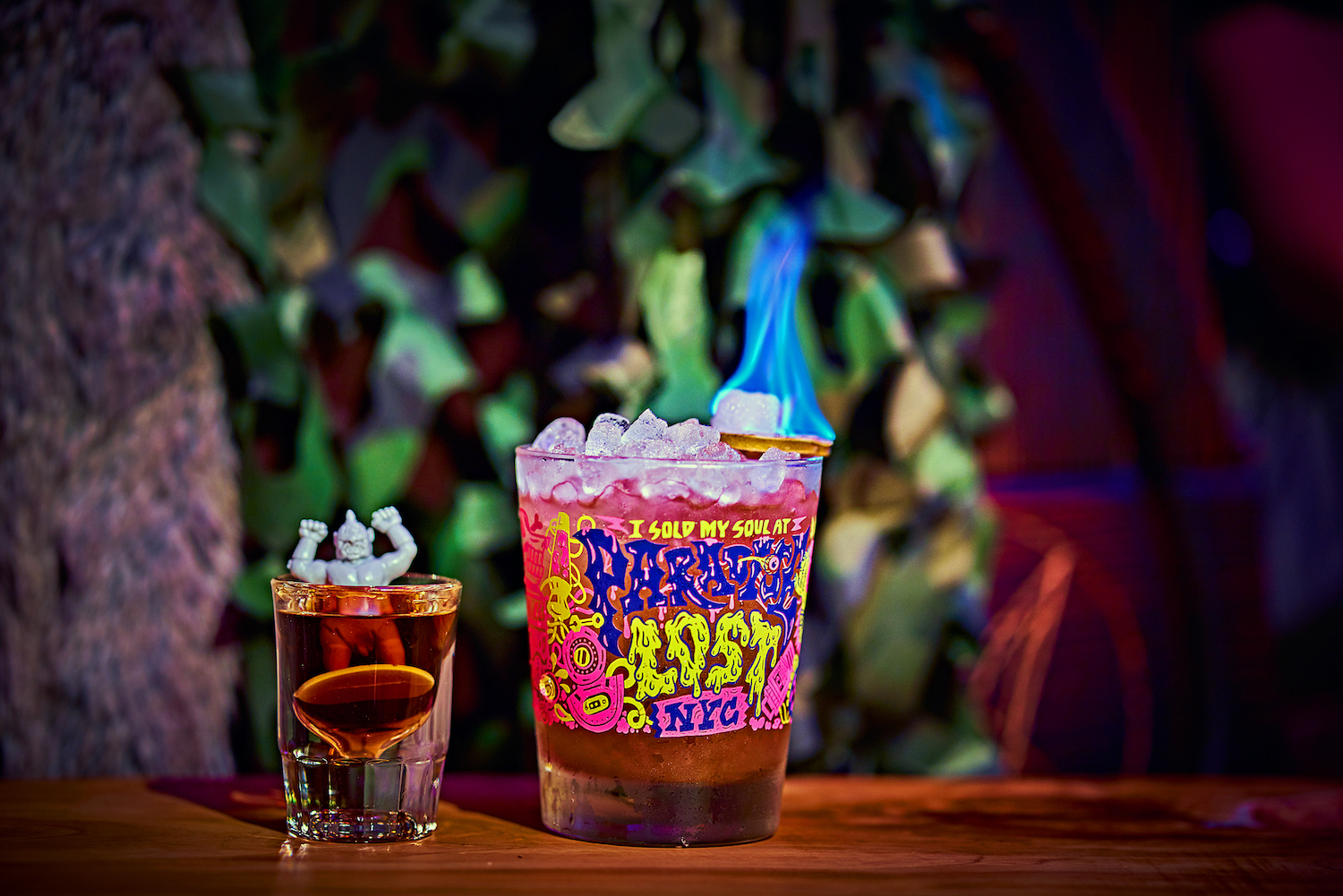 Cocktail in a lowball glass on fire next to a small shot with a plastic figurine inside of it.