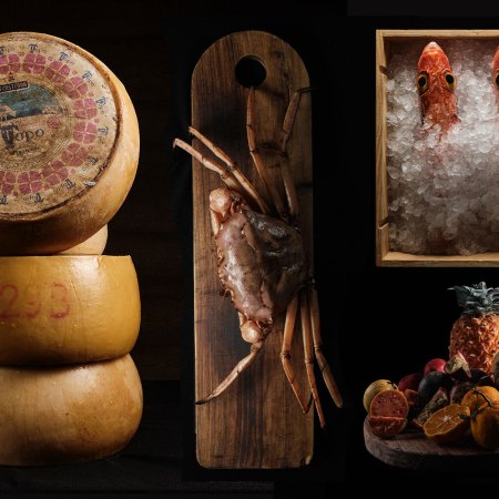 wheels of cheese, a crab, fish in a box of ice and a platter of tropical fruit on a black background