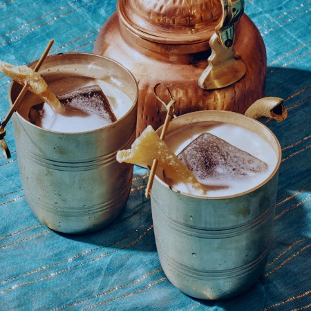 two cocktails in mule mugs garnished with candied ginger next to a teapot on a blue and gold tablecloth