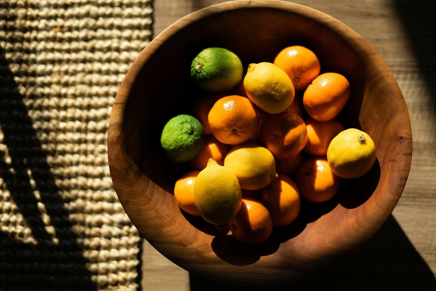 limes, oranges and lemons in a wooden bowl sitting next to a placemat on a wooden table