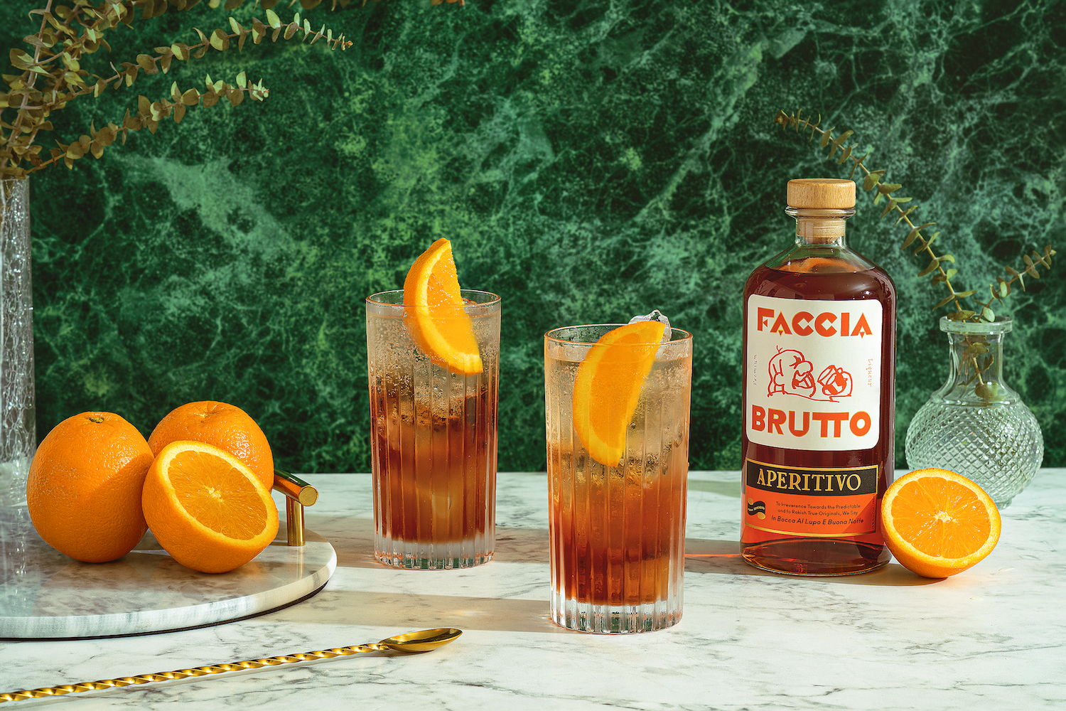 faccia brutto aperitivo next to 2 spritzes on a white marble table with a green marble background