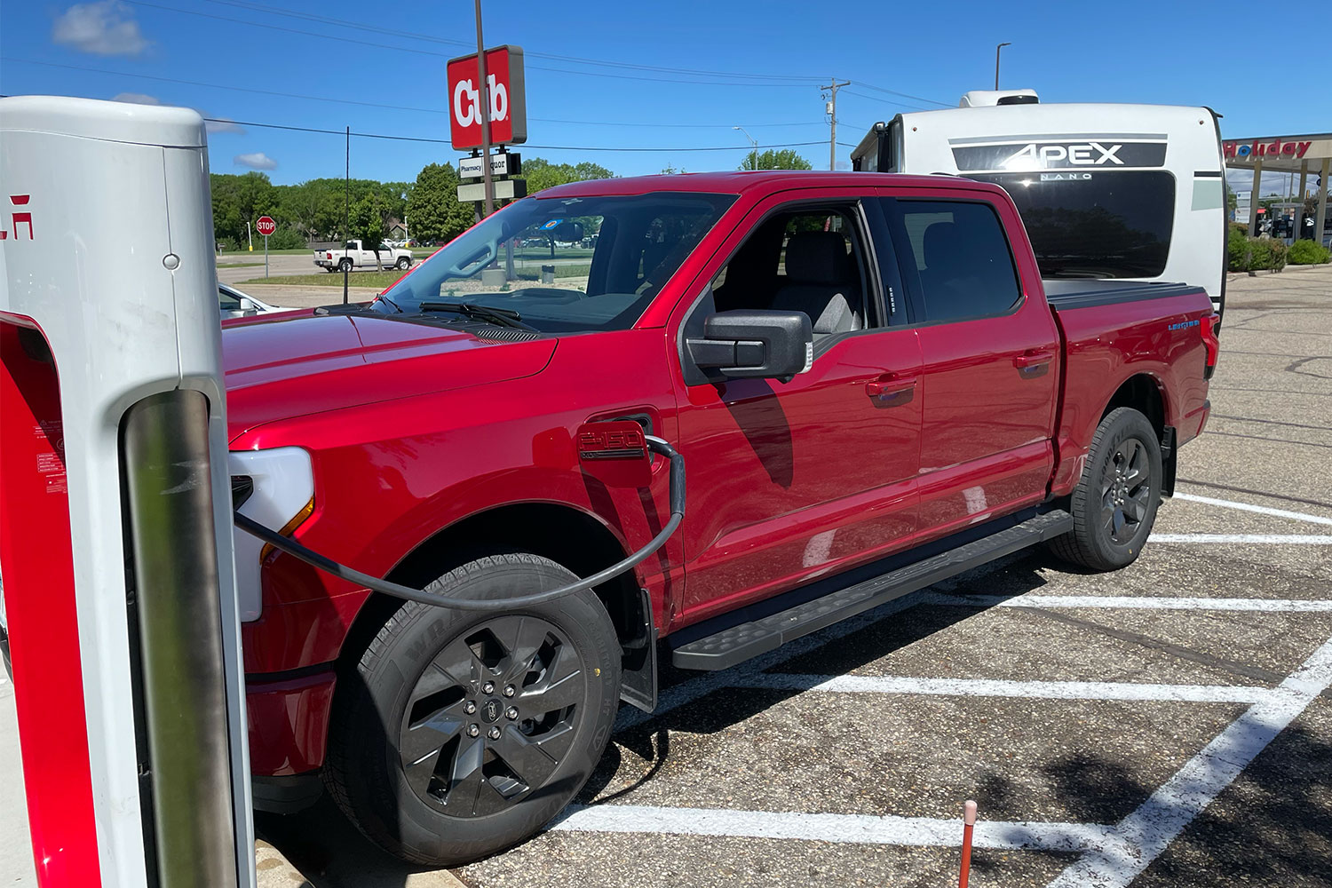 A red Ford F-150 Lightning electric pickup with a camper attached charging at a Tesla station