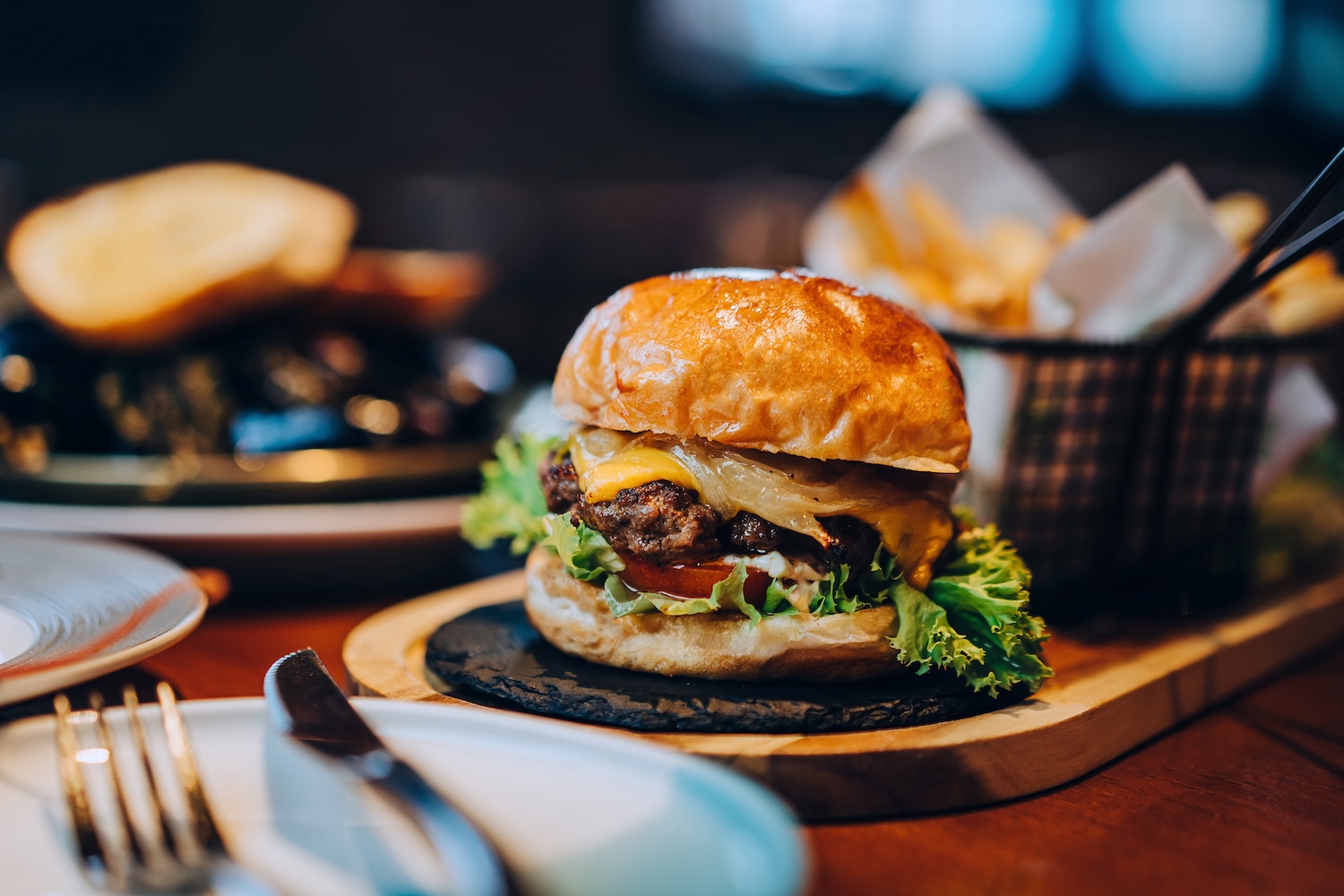 Fresh delicious burger with french fries and salad served on rustic wooden board on table. A feast on the dining table in restaurant. Food and lifestyle concept
