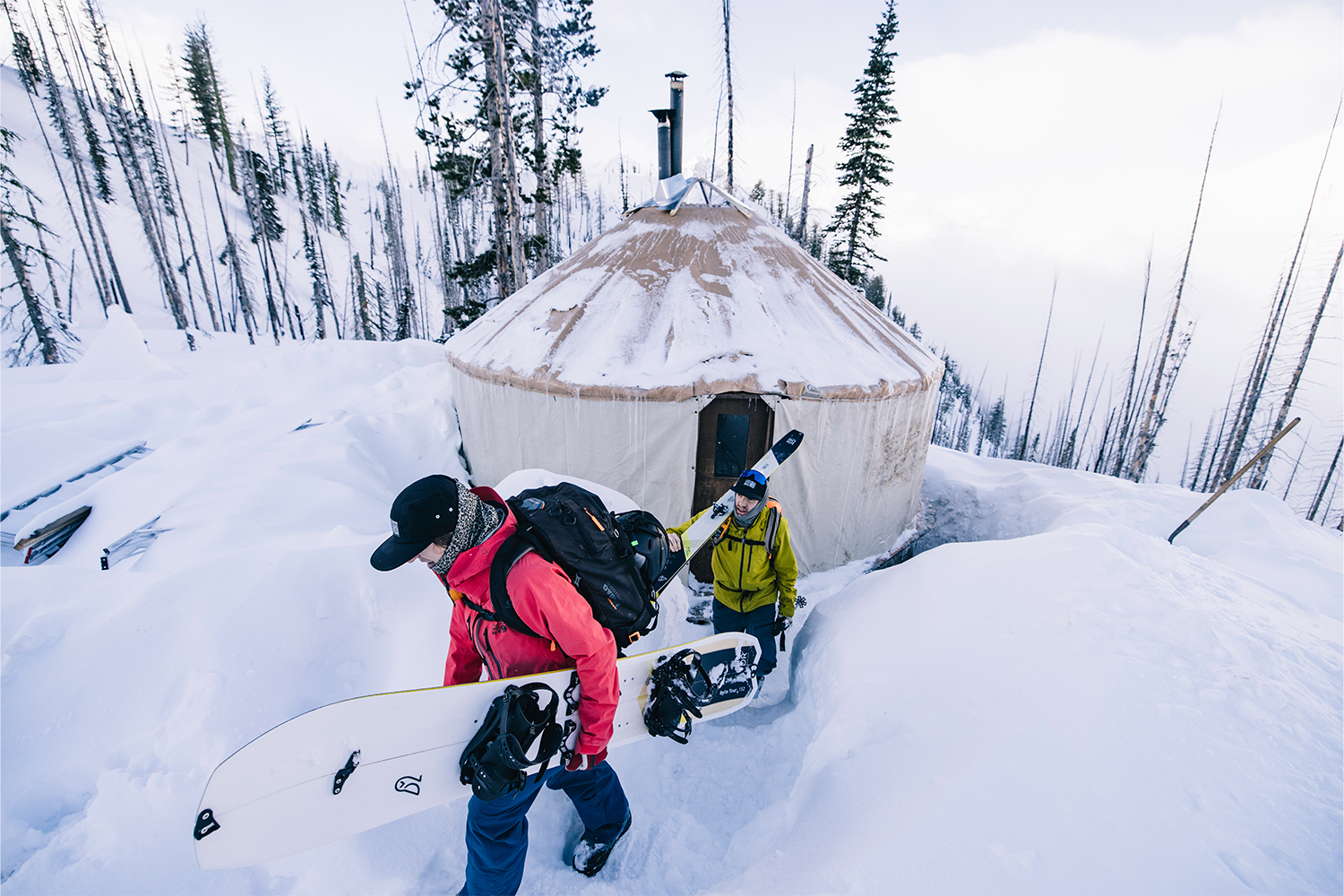 A pair of skiers depart the yurt and trudge through the snow with their gear.