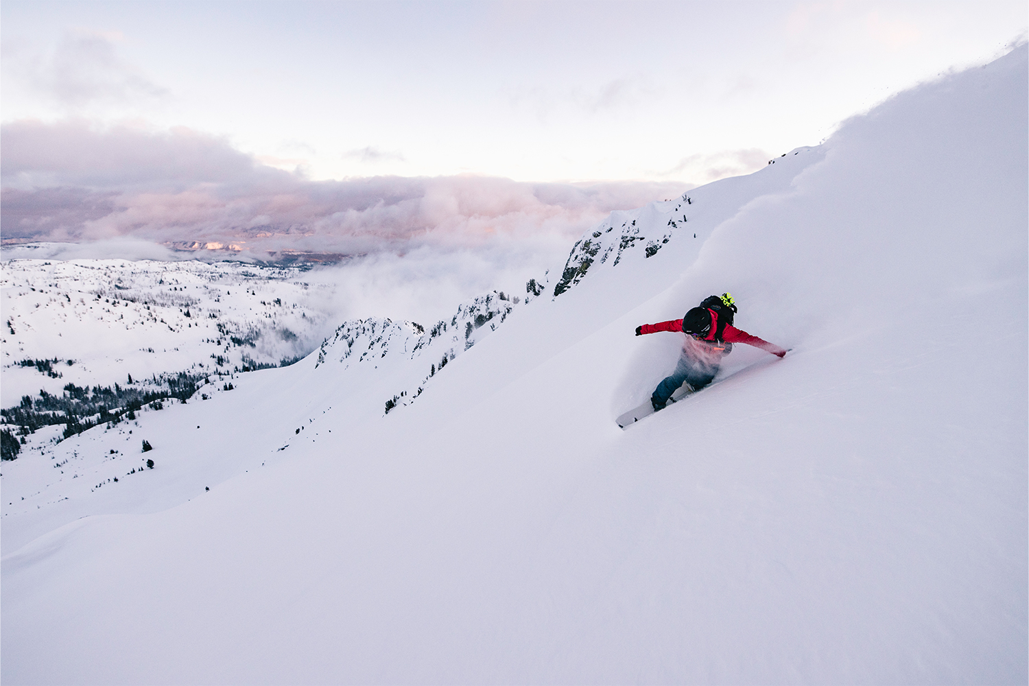A view of a snowboarder carving against the mountain, with purple clouds and white peaks in the background.