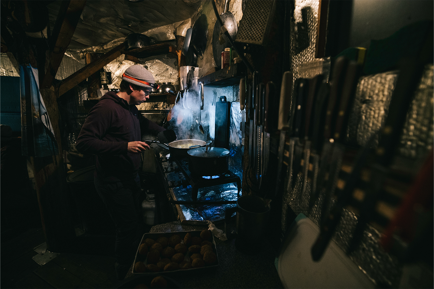 In the inside of the dark yurt, a skier cooks with a pan.