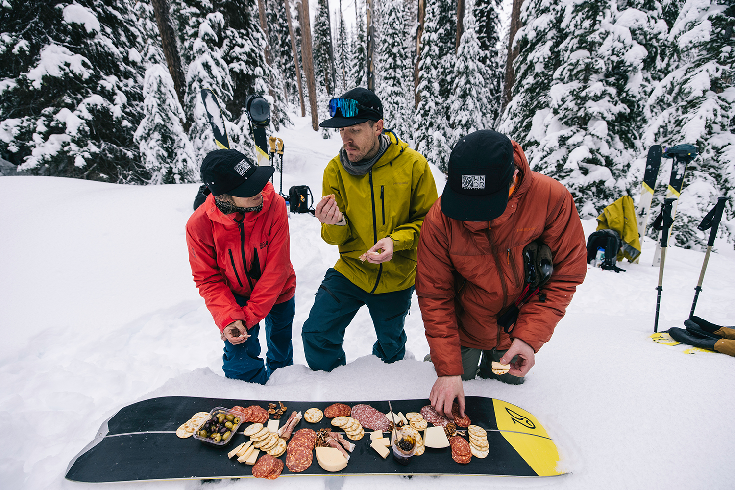 A trio eat charcuterie off a snowboard.