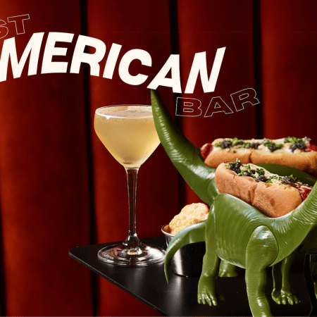 A cocktail from Yacht Club in Denver next to a dinosaur taco holder in front of a red velvet curtain