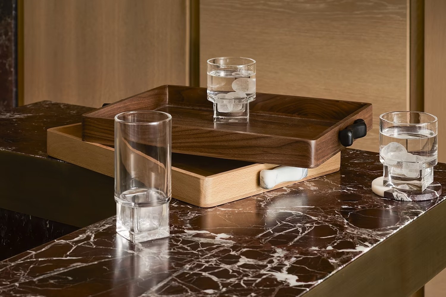 Three glasses, two filled with ice water, on stacked wooden trays and a dark marble counter.