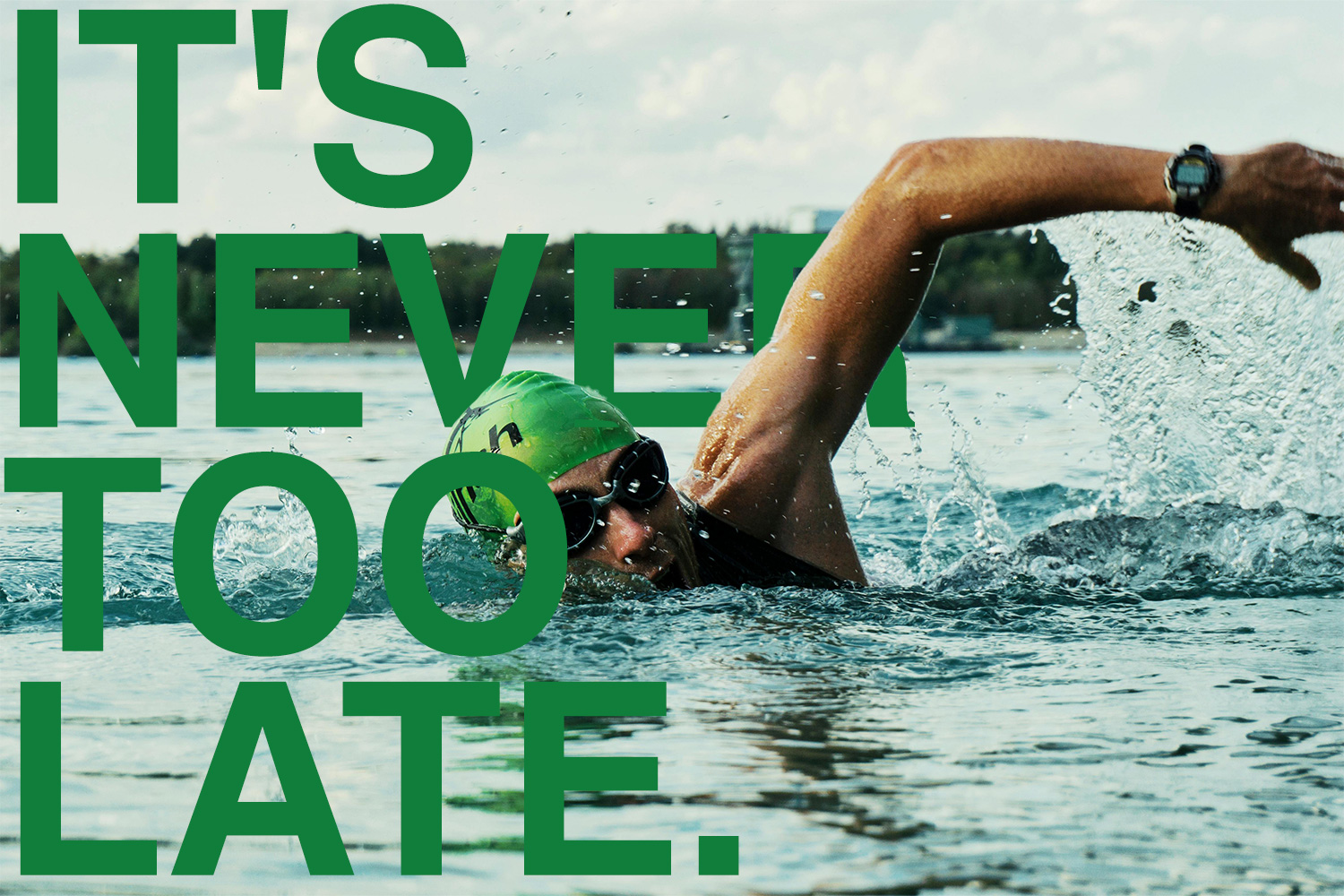Adult swimmer wearing green swim cap and goggles doing freestyle stroke in open water, overlaid with bold green text reading "It’s Never Too Late."