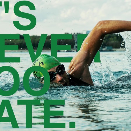 Adult swimmer wearing green swim cap and goggles doing freestyle stroke in open water, overlaid with bold green text reading "It’s Never Too Late."