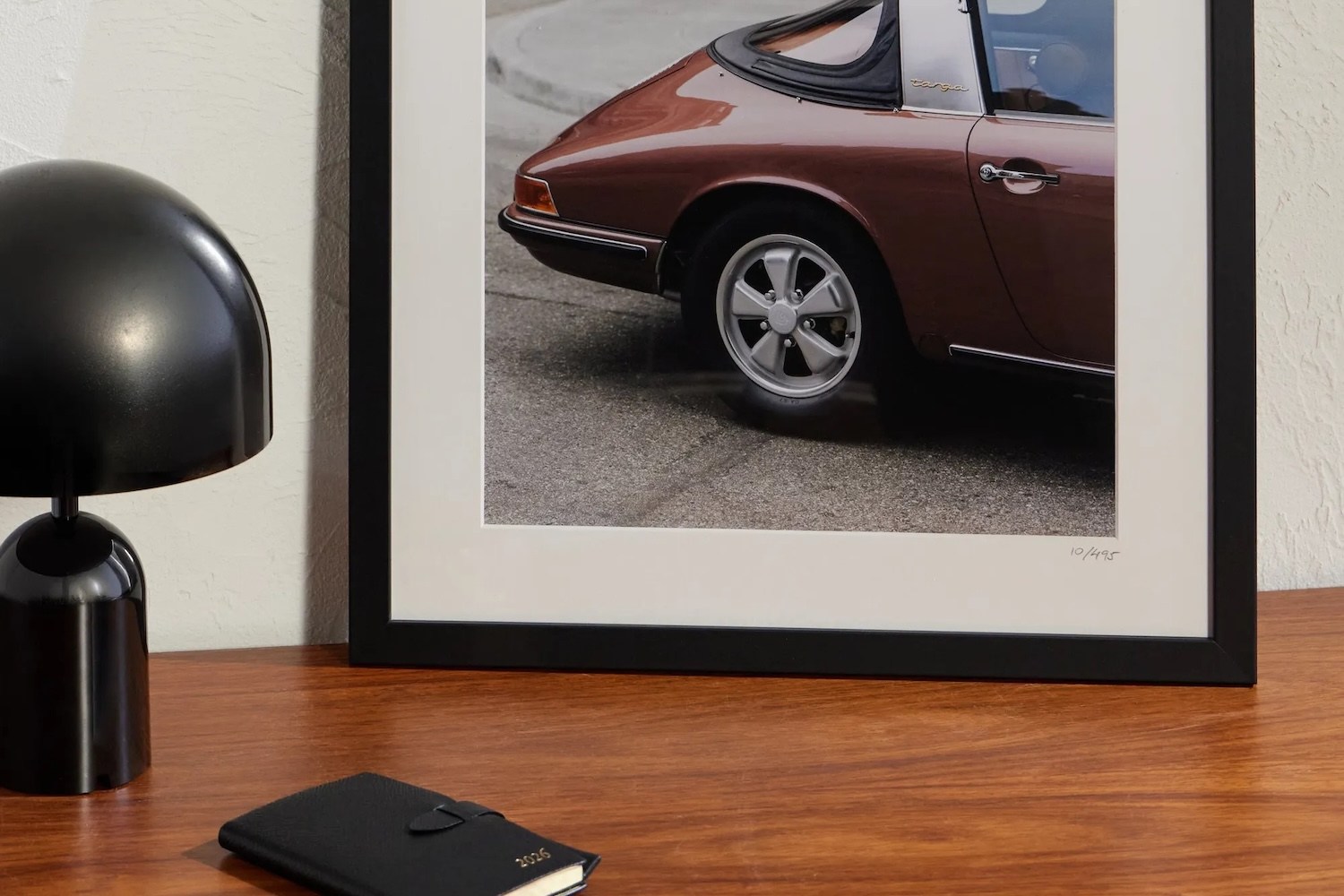 A framed print of a brown Porsche Targa, a black lamp, and a black 2026 planner on a wooden table.