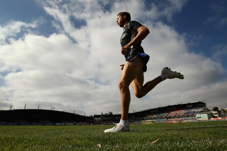 A male athlete running on a grass field in a stadium under a partly cloudy sky, wearing a black training outfit.
