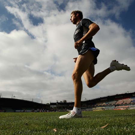 A male athlete running on a grass field in a stadium under a partly cloudy sky, wearing a black training outfit.