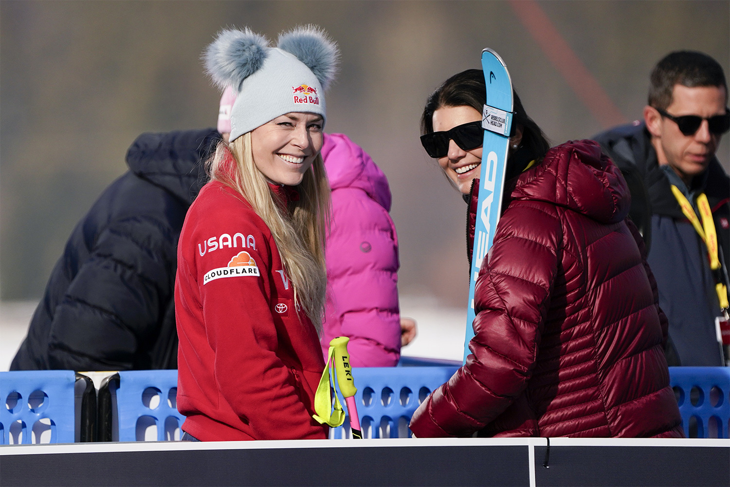 Lindsey Vonn smiles in a red jacket and beanie at a ski event, holding ski poles alongside fellow athletes.
