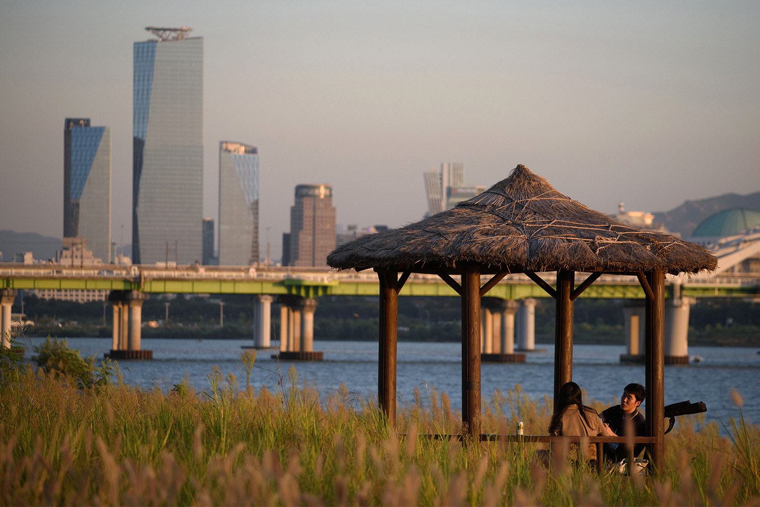 A young couple enjoys a quiet moment under a thatched-roof gazebo by the Han River in Seoul, with the modern skyline in the background, embodying the philosophy of sohwakhaeng—finding happiness in small, simple moments.
