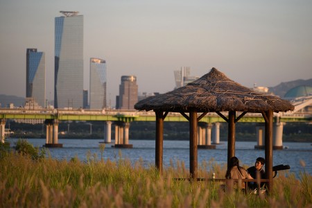 A young couple enjoys a quiet moment under a thatched-roof gazebo by the Han River in Seoul, with the modern skyline in the background, embodying the philosophy of sohwakhaeng—finding happiness in small, simple moments.