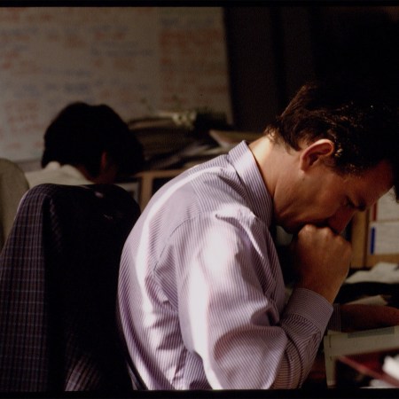 A stressed office worker in a striped dress shirt leans forward, deep in thought, with a cluttered desk and whiteboard in the background.