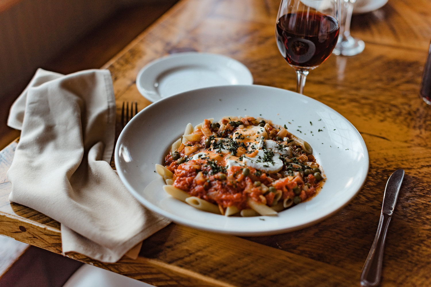 a bowl of red sauce pasta in a white bowl next to a glass of red wine on a wooden table