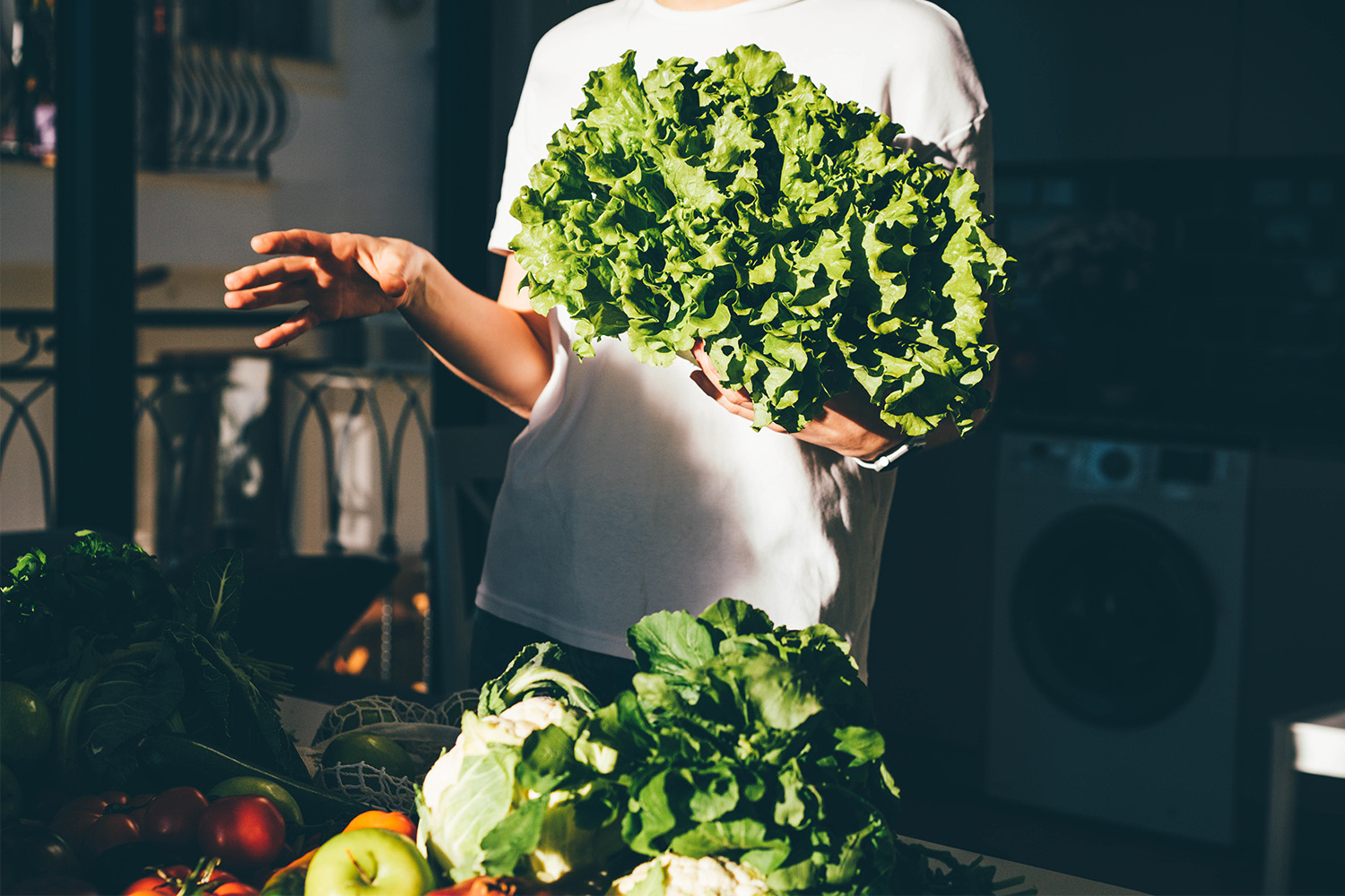 Person holding a large bunch of leafy greens, surrounded by fresh vegetables on a kitchen counter