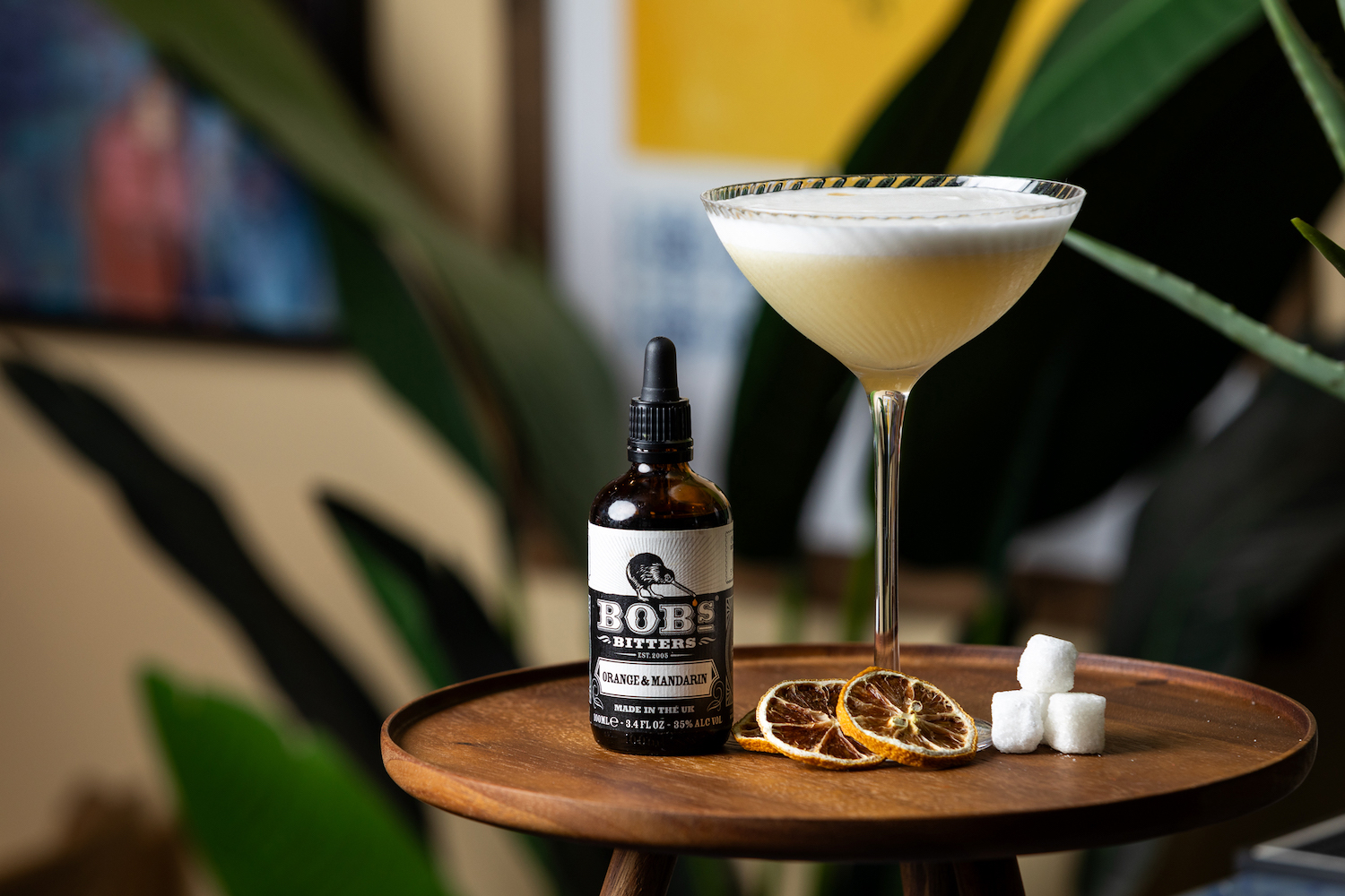 a bottles of bob's orange and mandarin bitters next to a sour cocktail in a coupe glass on a wooden table