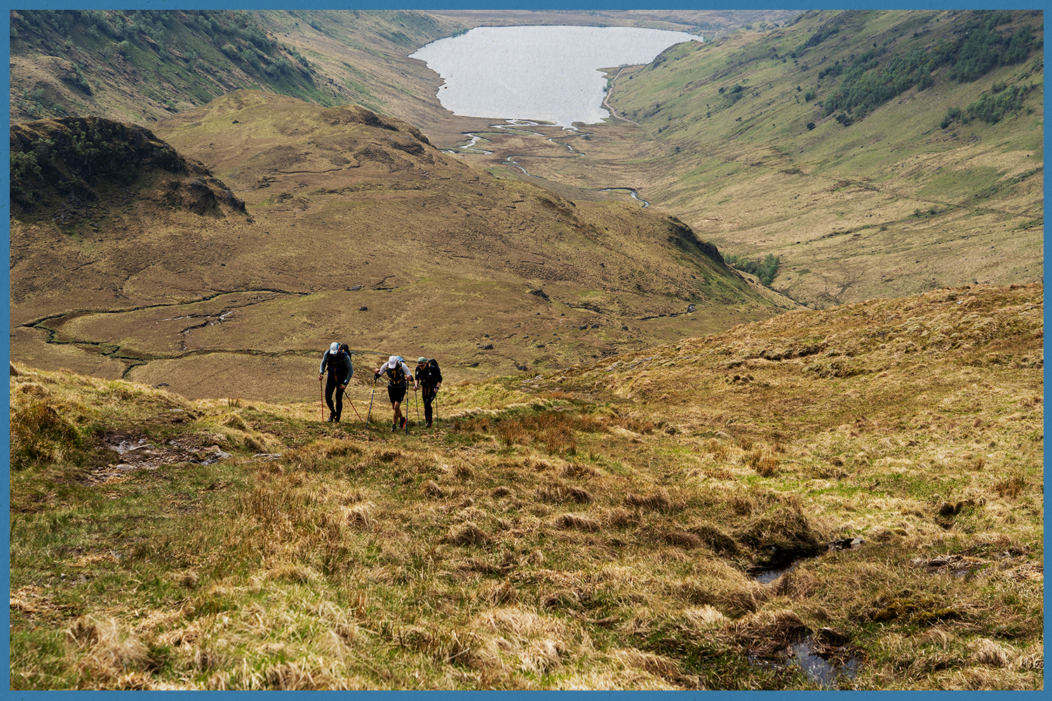 Three Highland Ultra runners ascend a steep hill overlooking a loch.