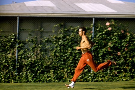Shirtless male runner in red track pants sprinting on grass with gauze around his feet.