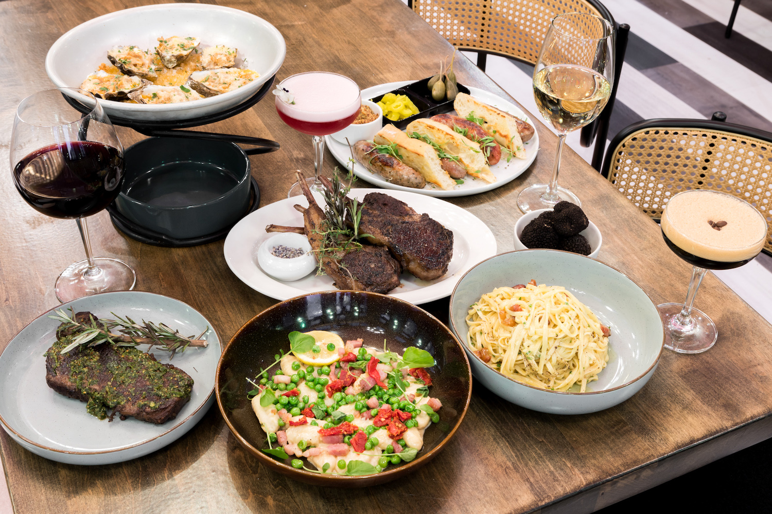 a spread of food on a wooden table with pasta, steak, lamb chops, baked oysters and an espresso martini