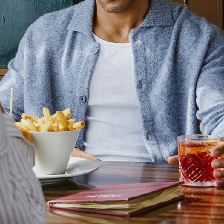 A person in a blue cardigan and white t-shirt holds a drink, with fries and a menu on the table.