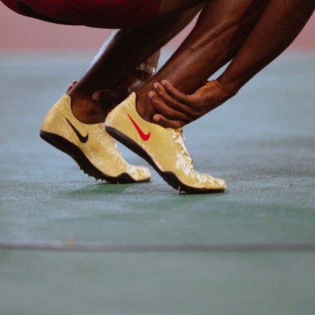 Close-up of powerful ankles in motion during a dynamic warm-up, wearing gold Nike spikes on a track surface.