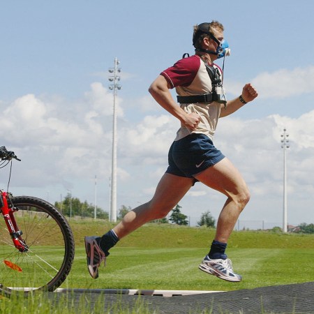 Man running with VO2 max mask on outdoor track next to coach on bike, symbolizing modern fitness optimization culture