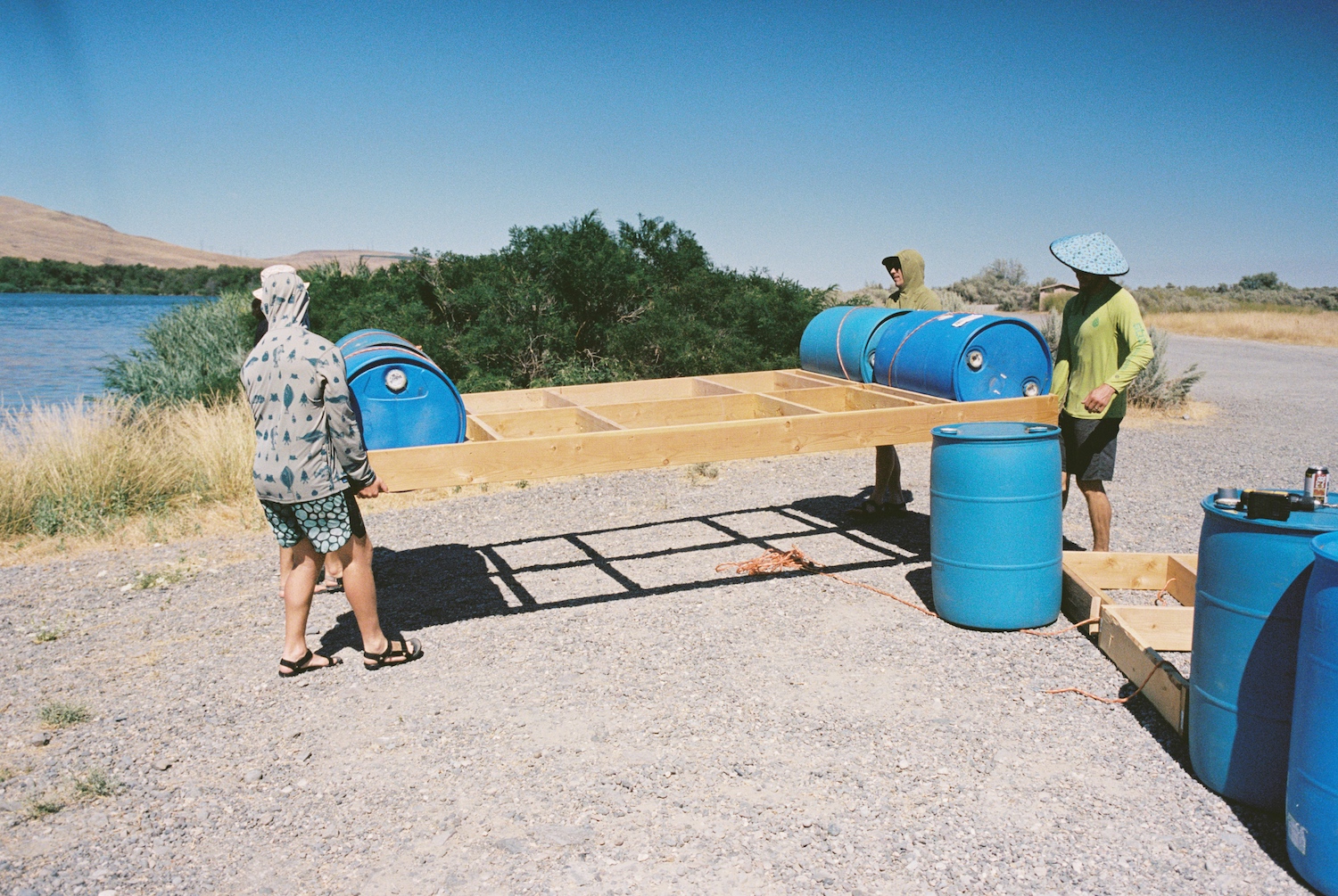 Three men carry pallets of wood and blue barrels along the river's edge.