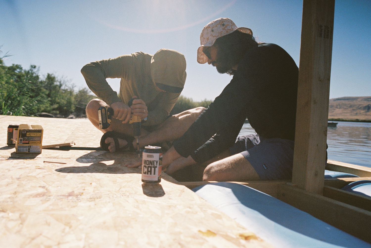 A pair of friends screw plywood into place while building the raft, with a beer can in the foreground.