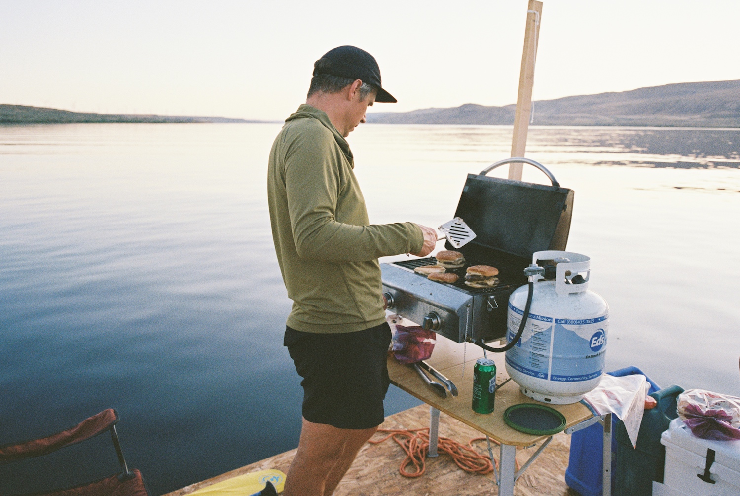 A man grilling burgers on a little grill aboard the raft, with a propane tank beside him.