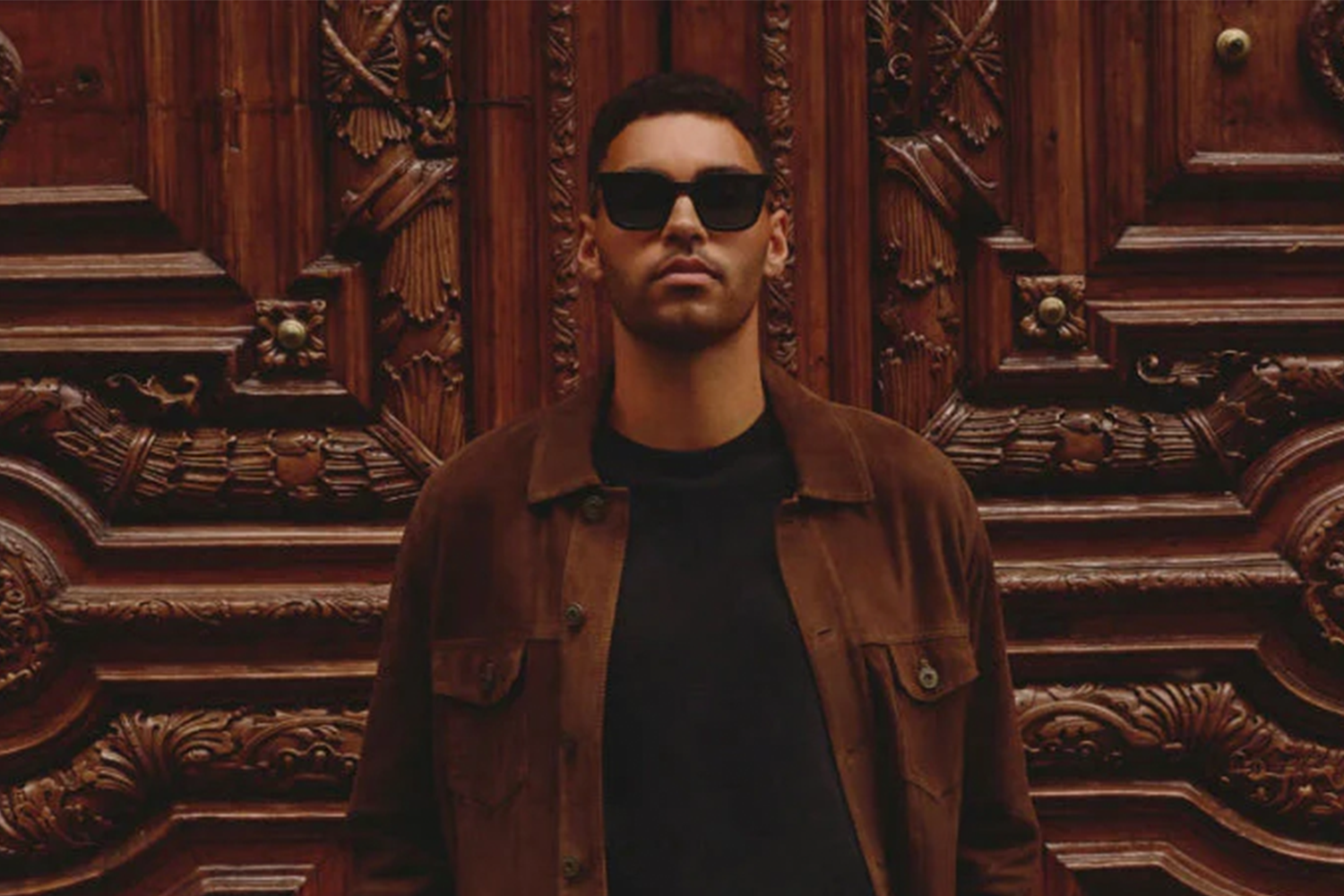 Man in sunglasses and a brown jacket standing in front of an ornate wooden door.