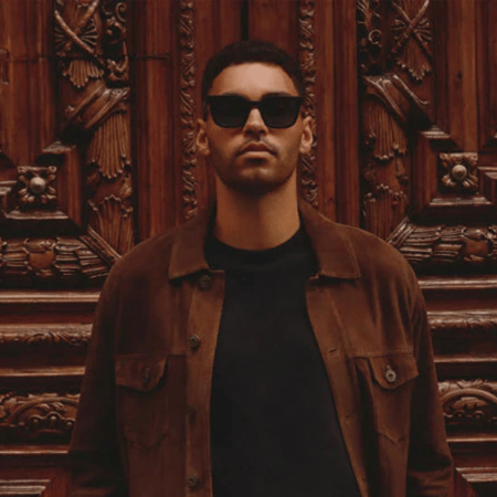 Man in sunglasses and a brown jacket standing in front of an ornate wooden door.