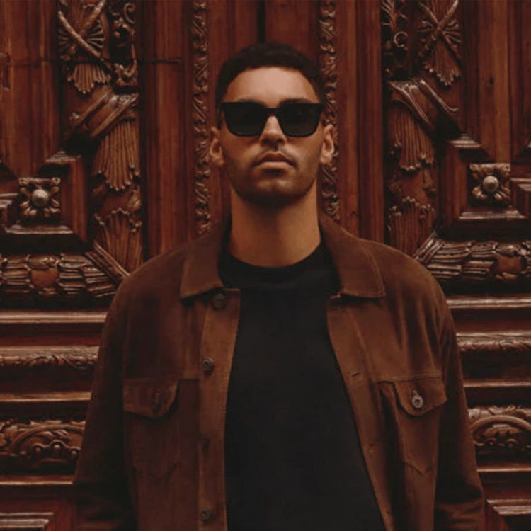 Man in sunglasses and a brown jacket standing in front of an ornate wooden door.