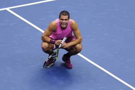 Carlos Alcaraz smiles while crouching on a tennis court.