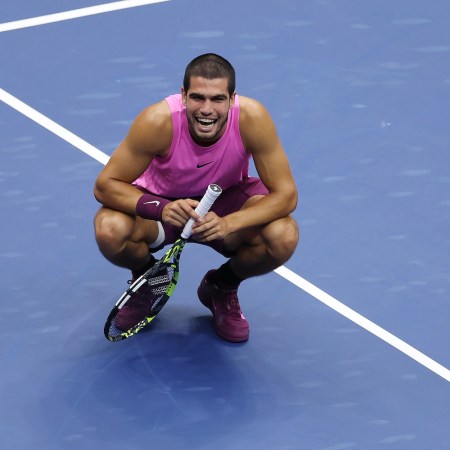 Carlos Alcaraz smiles while crouching on a tennis court.