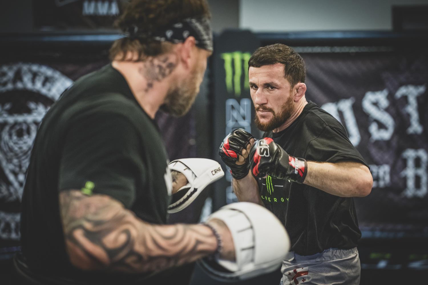 An MMA fighter squares up against a coach with his fists up. The coach with his back turned is wearing white focus mitts.