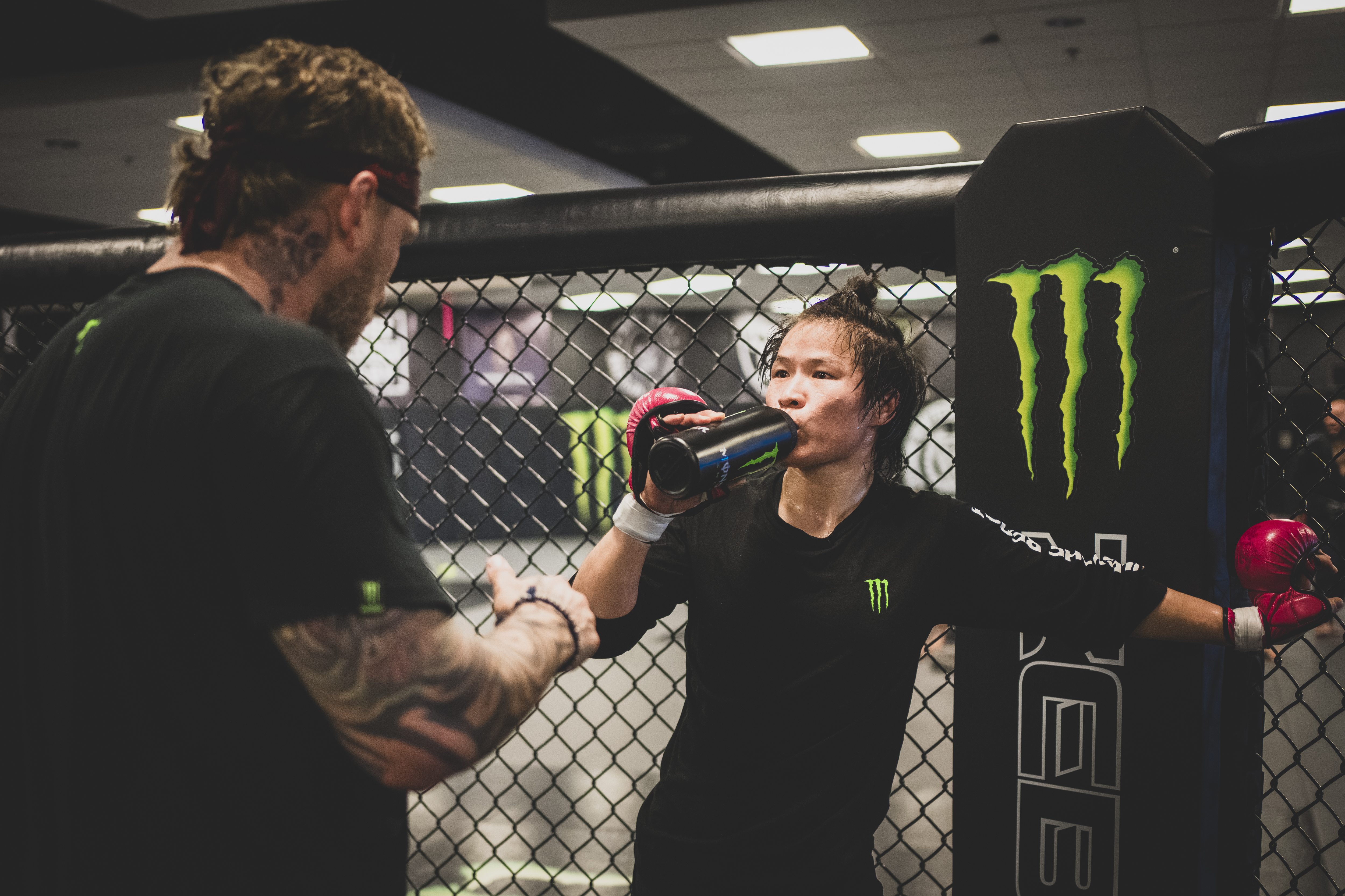 A female MMA fighter drinks from a water bottle while wearing fighting gloves and talking to her coach