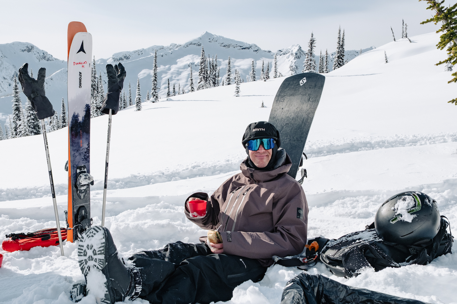 A man leaning against a snowboard, sipping coffee.